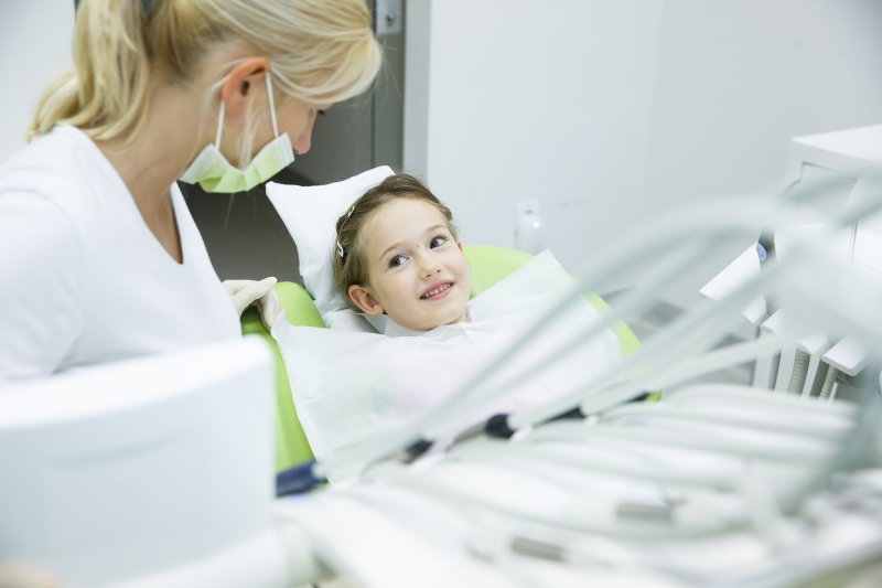 Dentist wearing mask talking to smiling child in green dental chair with equipment in the foreground