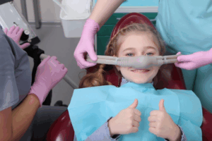 Child giving two thumbs up as dentist administers laughing gas